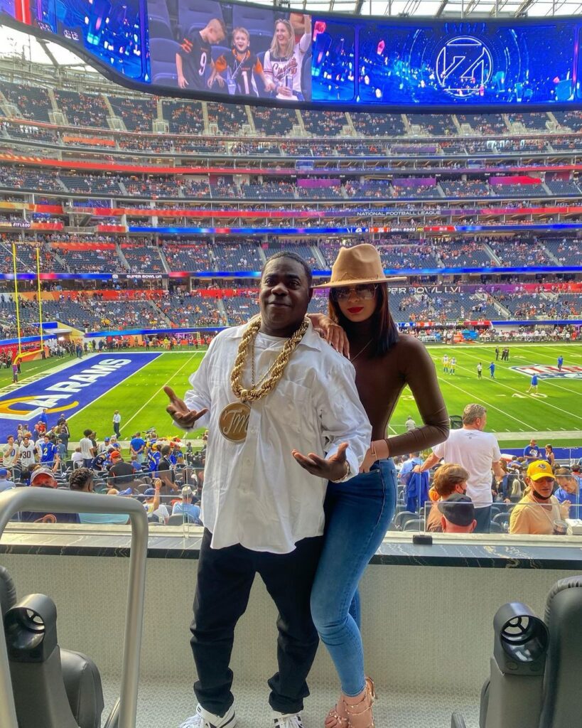 Tracy wearing white tee gold chain and black jeans and posing with his partner in a stadium.