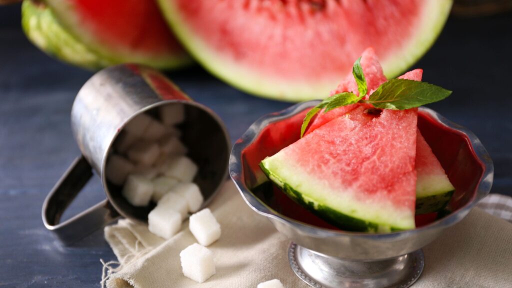 Watermelon served on a silver plate on the table. Also, a half cut watermelon is placed on the side along with a silver cup full of sugar cubes.
