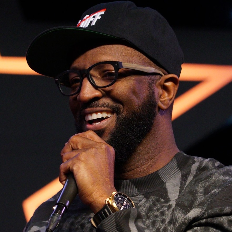 Rickey Smiley wearing black cap and printed tee and performing on stage.