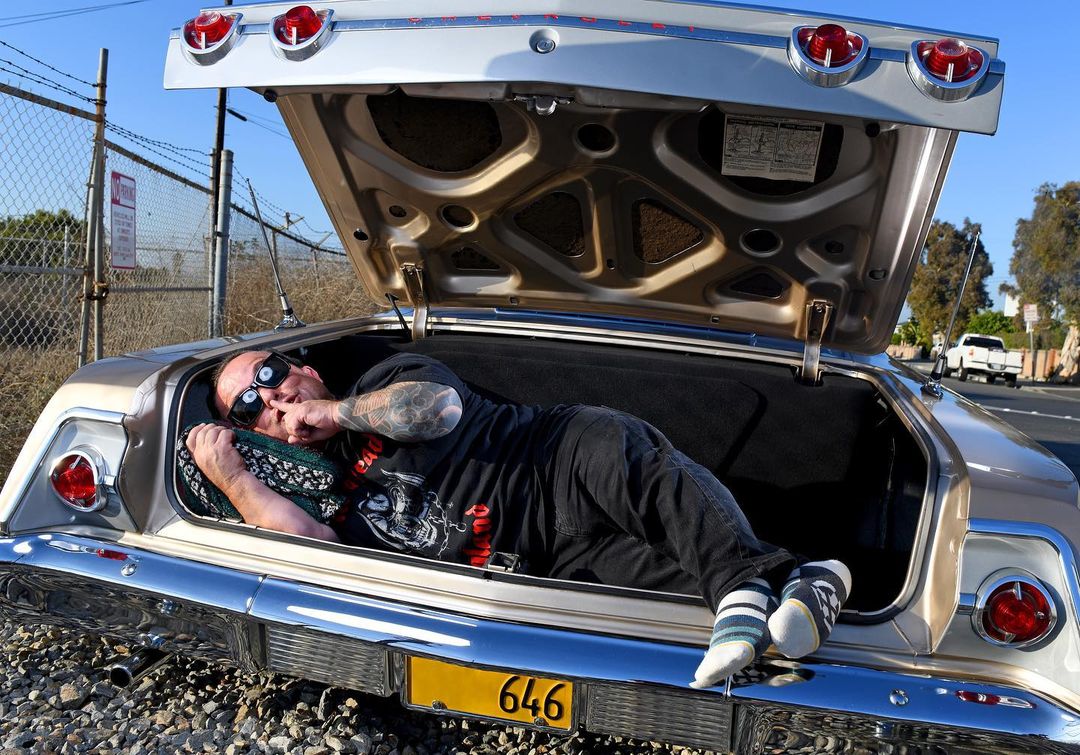 Wee Man sleeping on the back of his car. He is wearing sunglasses.