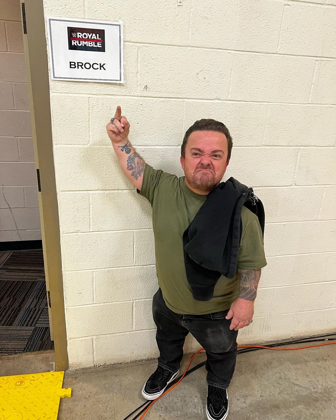 Wee Man wearing green shirt and blck pants pointing to a board on the side of the door.