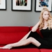 Sara Mitich wearing a black dress and posing on a couch.