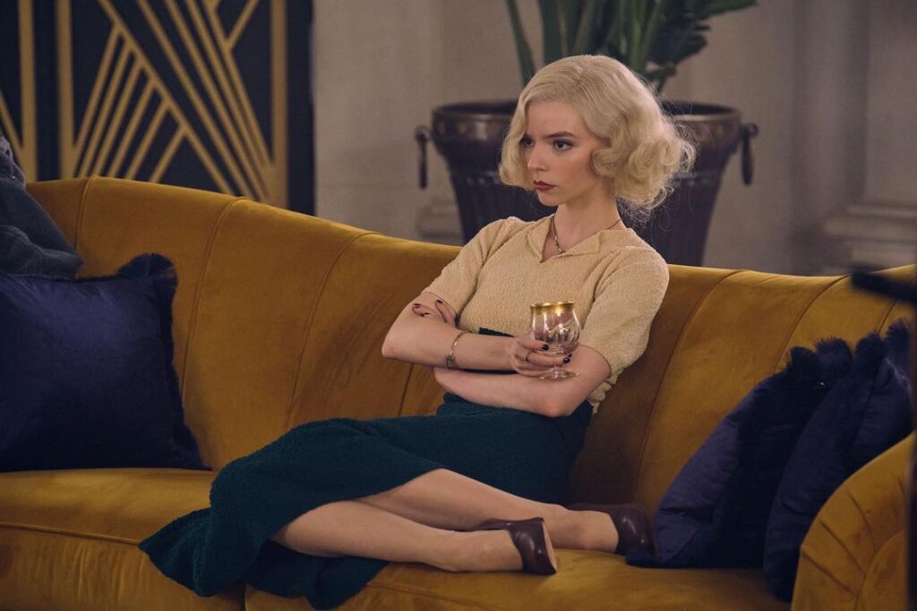 Anya Taylor-Joy sitting on a sofa wearing black skirt and cream top.